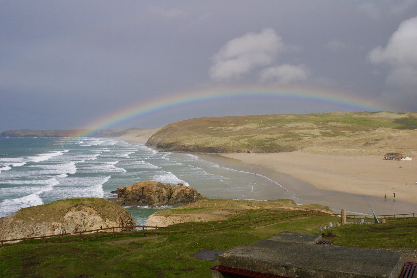 A rainbow over Perranporth beach with long lines of surf running along the bay