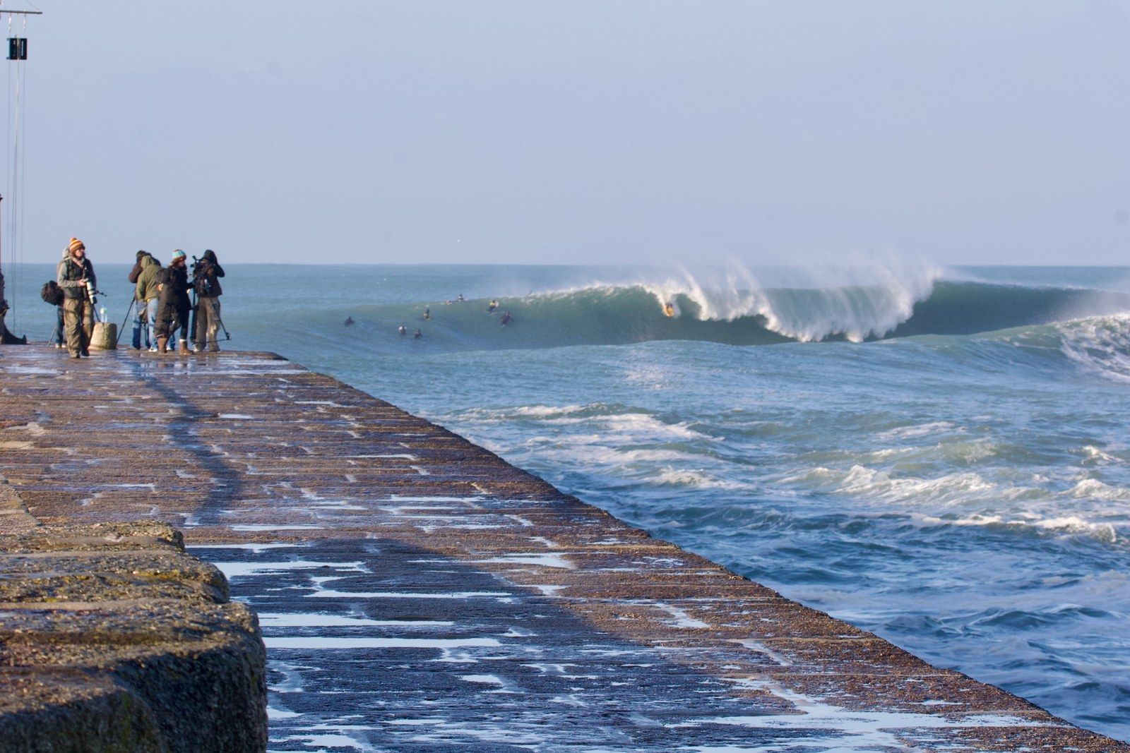 A clean Porthleven reef wave breaking beside the harbour wall, with surfers and photographers watching